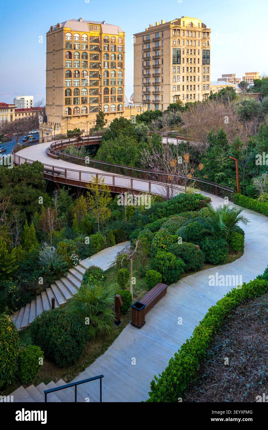 Passerella pedonale rialzata curva sopra i giardini paesaggistici nel parco Chambarakand con edifici residenziali Baku alle spalle. Baku, Azerbaigian. Foto Stock