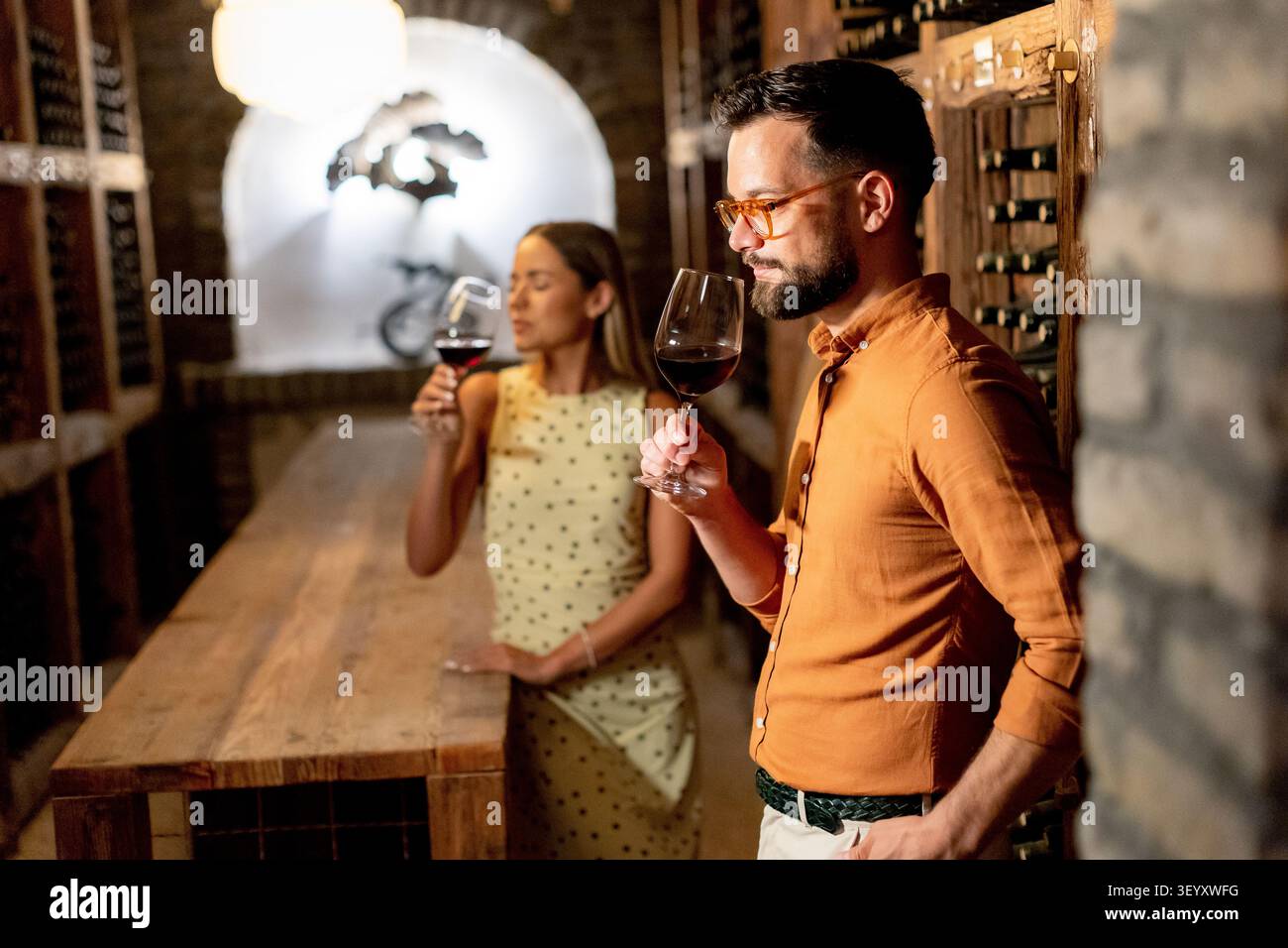 Un uomo e una donna sono in piedi in una cantina con dei bicchieri di vino rosso. Assaggiano il vino mentre si godono l'atmosfera. L'illuminazione lo è Foto Stock