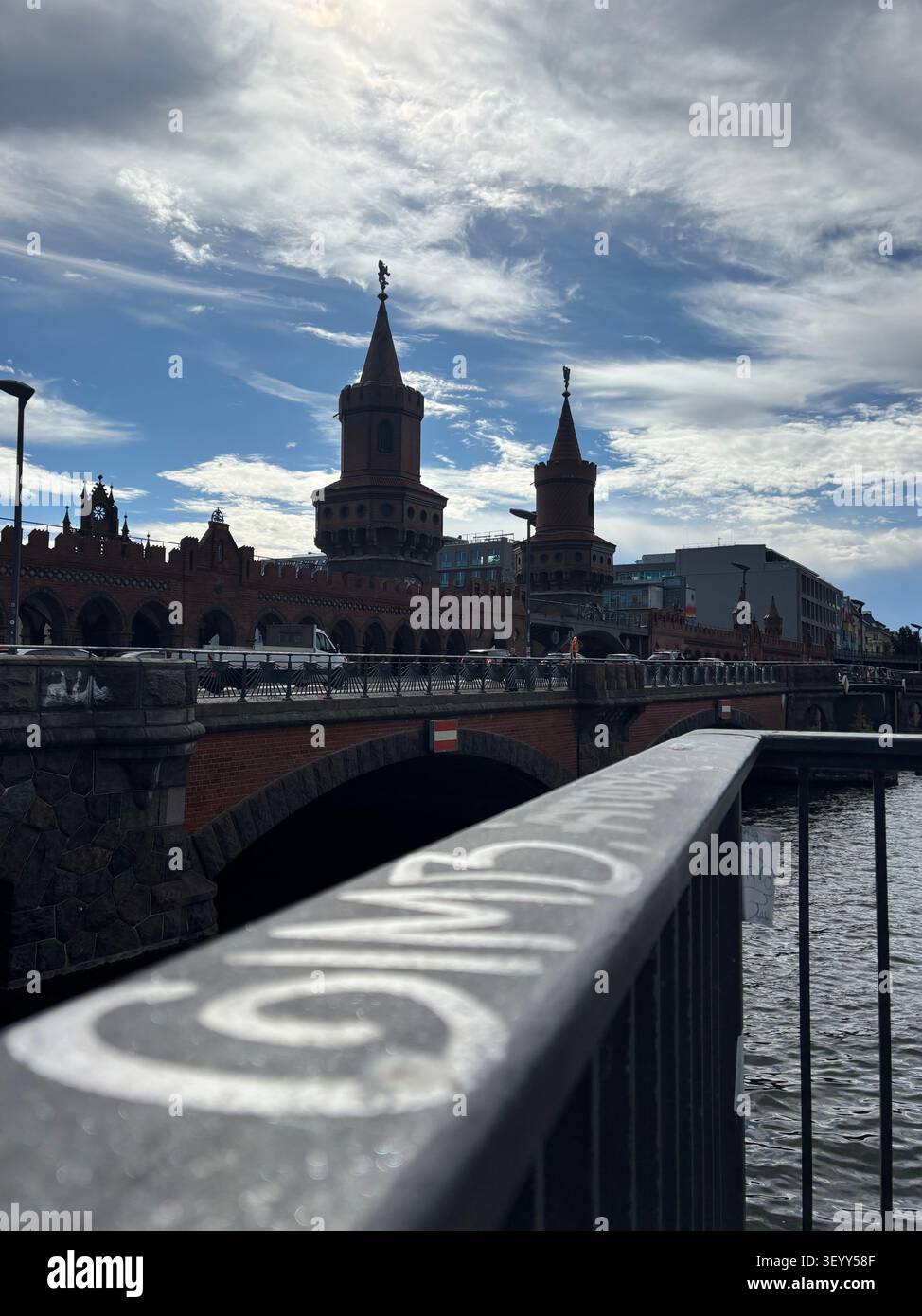 Ampia prospettiva del ponte storico in mattoni Oberbaum che si estende sul fiume Spree sotto un cielo drammatico, incorniciata da una ringhiera con graffiti in primo piano, Berlino - Immagine stock catturata con smartphone