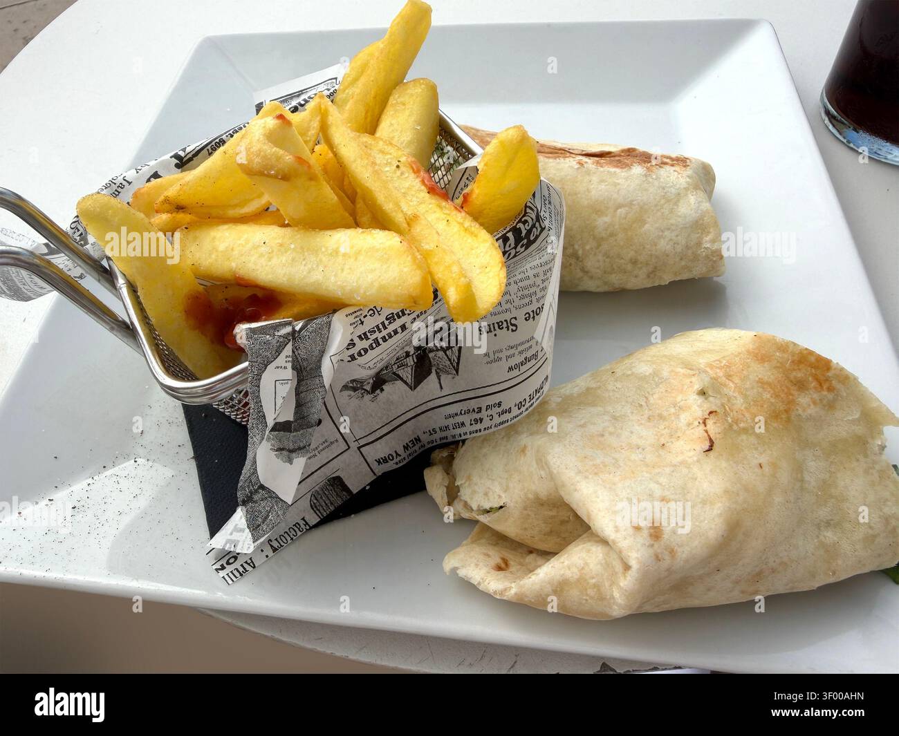 Un piatto bianco servito con due involtini di tortilla e patatine fritte dorate in un piccolo cestino di fili foderato con stampa di giornali finti - Immagine stock catturata con smartphone