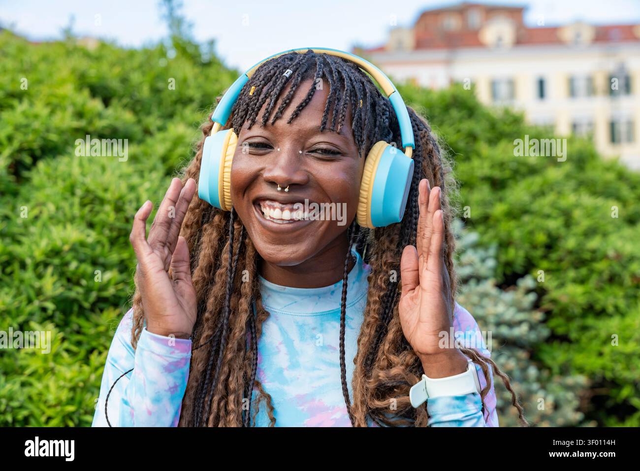 Ritratto ravvicinato di una giovane donna sorridente con capelli intrecciati che indossa cuffie sovrauricolari colorate all'aperto, tenendo i padiglioni mentre ascolti i mu Foto Stock