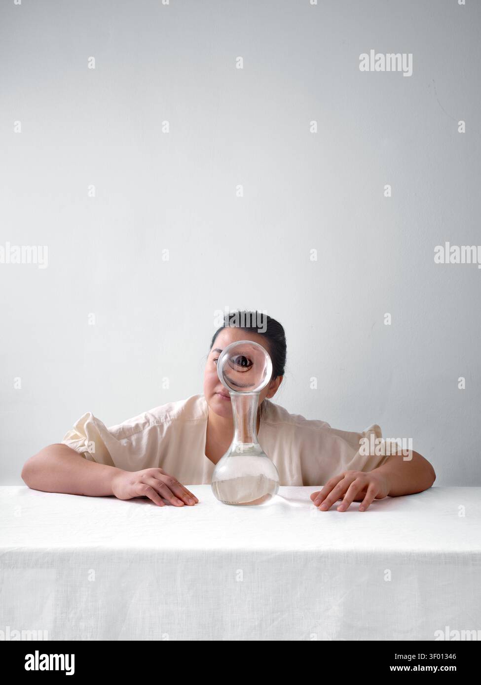 Ritratto minimalista di una donna seduta a un tavolo, che guarda attraverso una sfera di cristallo trasparente che ingrandisce il suo occhio, creando un concetto surreale di visione Foto Stock