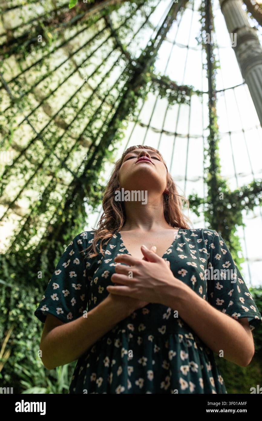 Ritratto ad angolo basso di una donna con le mani sul petto, gli occhi chiusi, respirando profondamente sotto una serra di vetro ricoperta di vite, sentendosi calmo e connesso Foto Stock