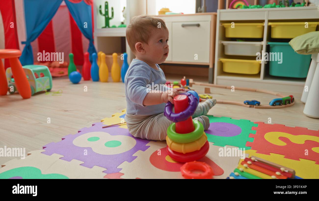 Bambino seduto su un tappetino in schiuma che tiene un anello viola con le mani e si impila un giocattolo nell'edificio; curiosità apprendimento sviluppo. Foto Stock