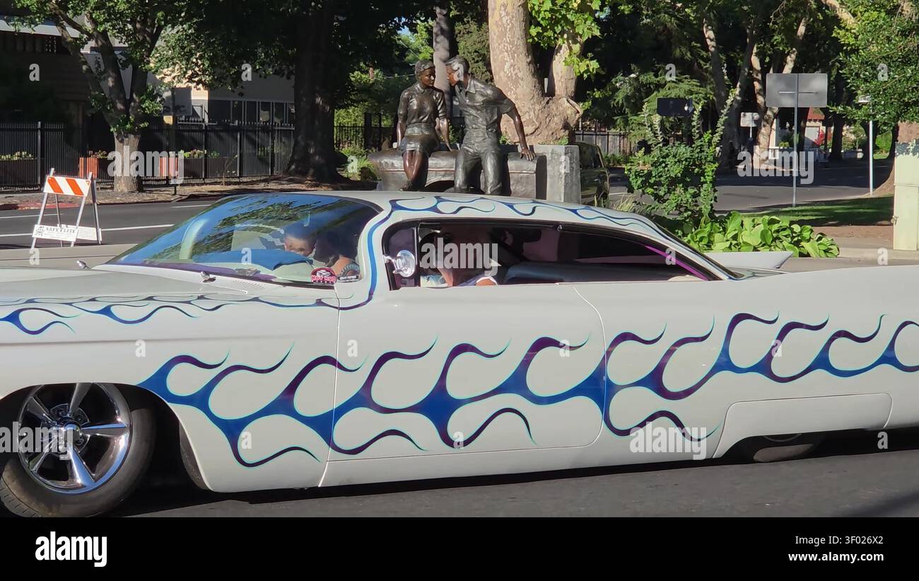 Un'auto d'epoca personalizzata con verniciatura a fiamma blu che guida lungo una strada, passando davanti a una scultura in bronzo di una coppia. Foto Stock