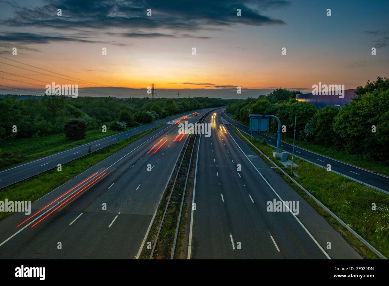 Lunga esposizione di percorsi leggeri per veicoli su un'autostrada M4 a più corsie al tramonto con un cielo all'orizzonte arancione e alberi verdi, Regno Unito. 13 giugno Foto Stock