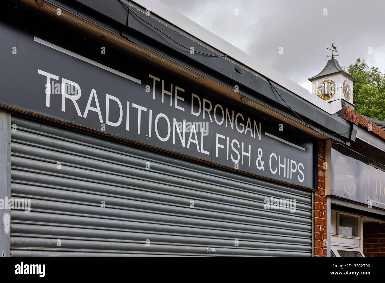 Drongan, Regno Unito - 24 maggio 2025: Il cartello per il tradizionale pesce e patatine Drongan sopra una serranda metallica chiusa, con la torre dell'orologio del negozio del villaggio Foto Stock