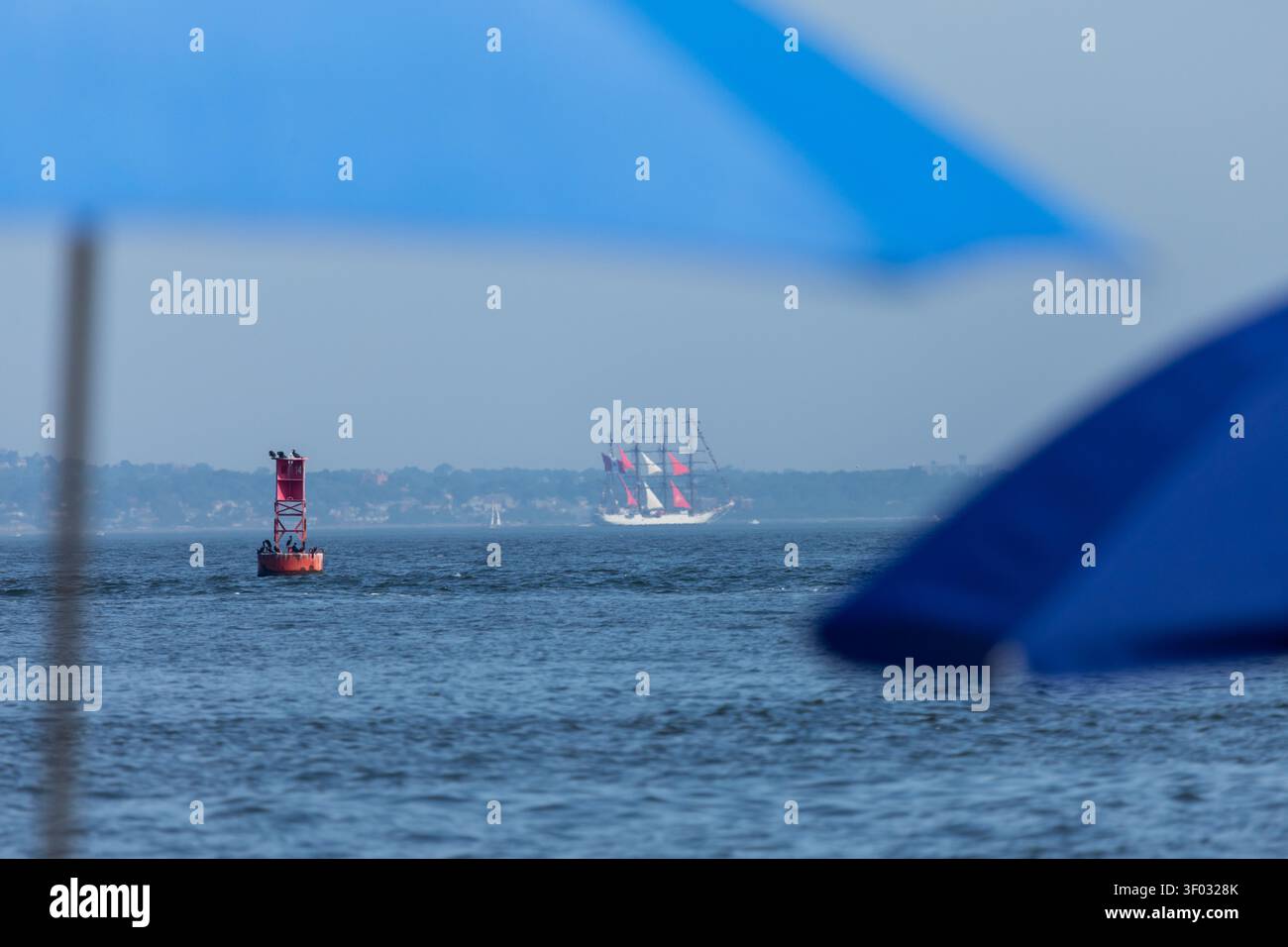 Sandy Hook, New Jersey, Stati Uniti d'America – 4 luglio 2026: Gli ombrelloni sfocati incorniciano un lontano veliero con vele rosse cerimoniali mentre naviga attraverso Sandy Hook Foto Stock