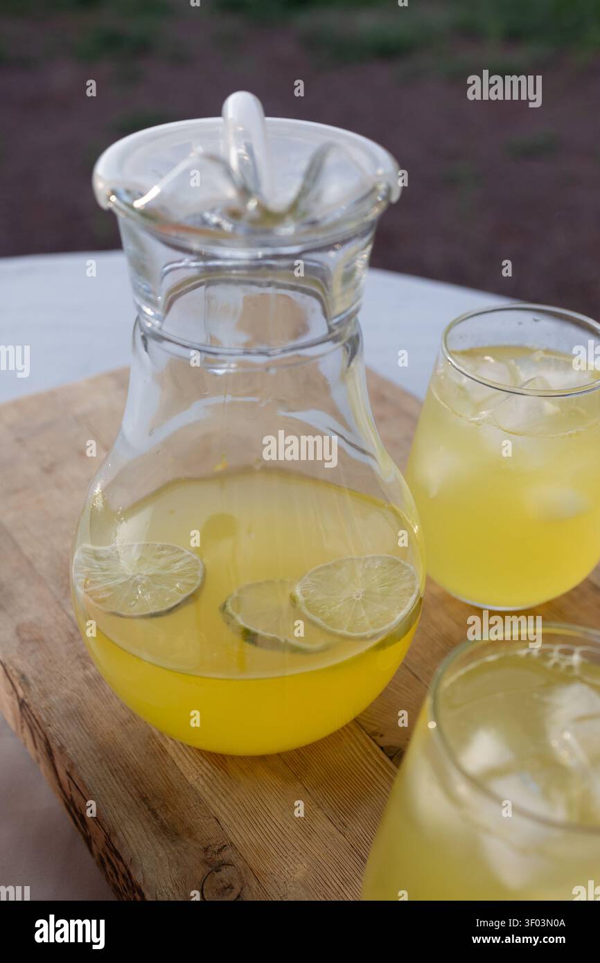Limonata servita in una caraffa e bicchieri con, in ambiente esterno eventualmente in occasione di un evento o di una riunione, con caraffa, bicchieri, cubetti di ghiaccio, fette di lime, con superficie in vetro e legno. Foto Stock