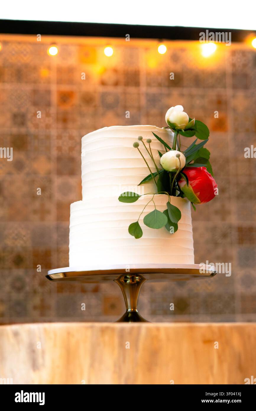 Una torta nuziale bianca a due piani con crema al burro strutturata è sormontata da fiori di peonia bianchi e bordeaux su un supporto d'argento con luci a filo dietro Foto Stock
