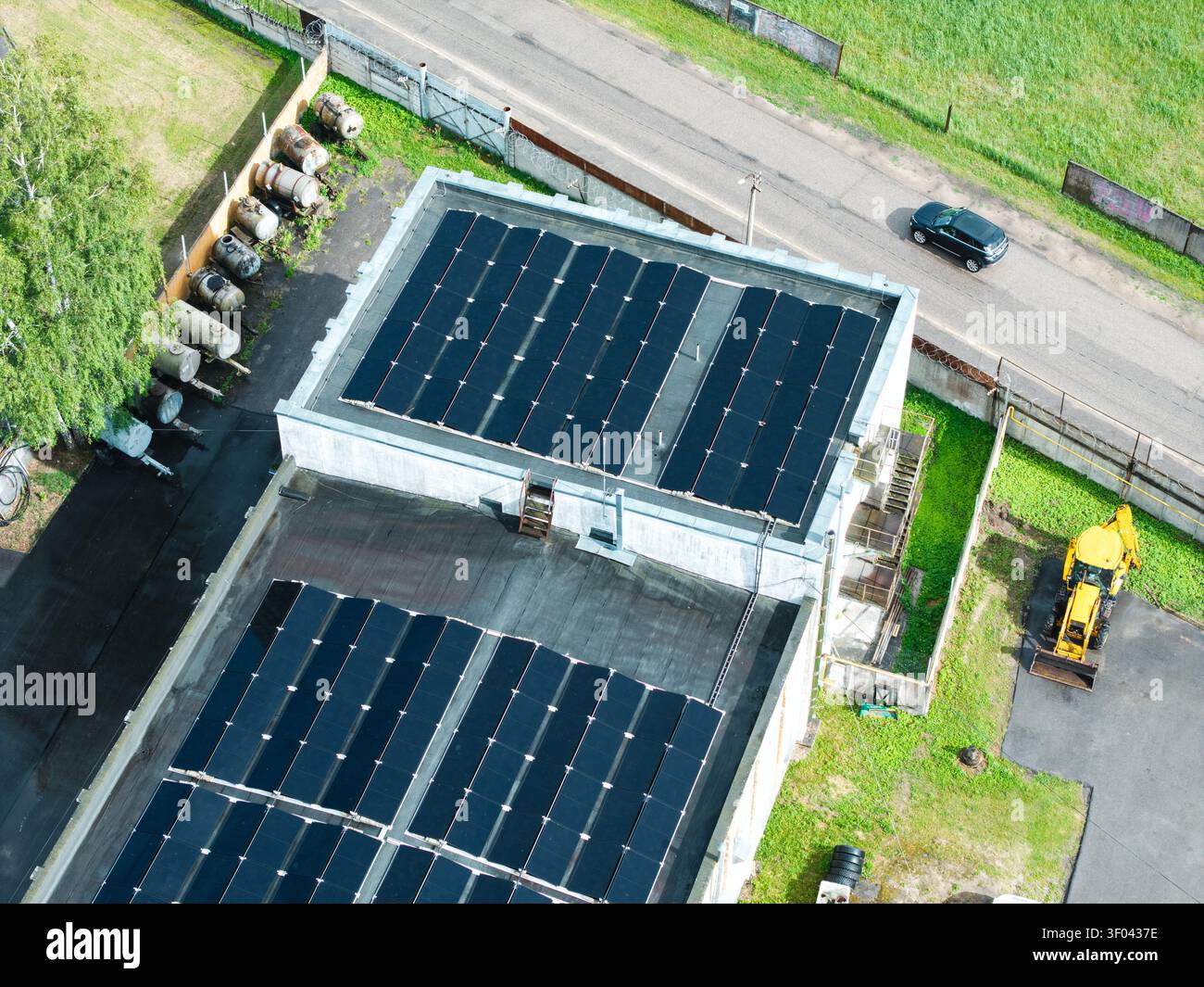 Vista aerea dei pannelli solari installati su un tetto piatto di un edificio industriale con una terna gialla parcheggiata nelle vicinanze e serbatoi di stoccaggio a Olaine, Olain Foto Stock
