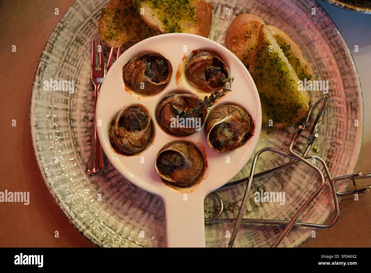 Vista dall'alto di lumache alla griglia cotte al forno in una pirofila di ceramica bianca con pane alle erbe e pinza da servizio. Cucina francese raffinata e presentazione elegante da ristorante. Foto Stock