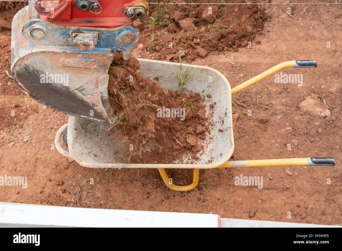 Primo piano dinamico di terreno marrone scuro che cade dalla benna di un escavatore in metallo in una carriola gialla. Acquisizione del processo attivo dei lavori in terra Foto Stock