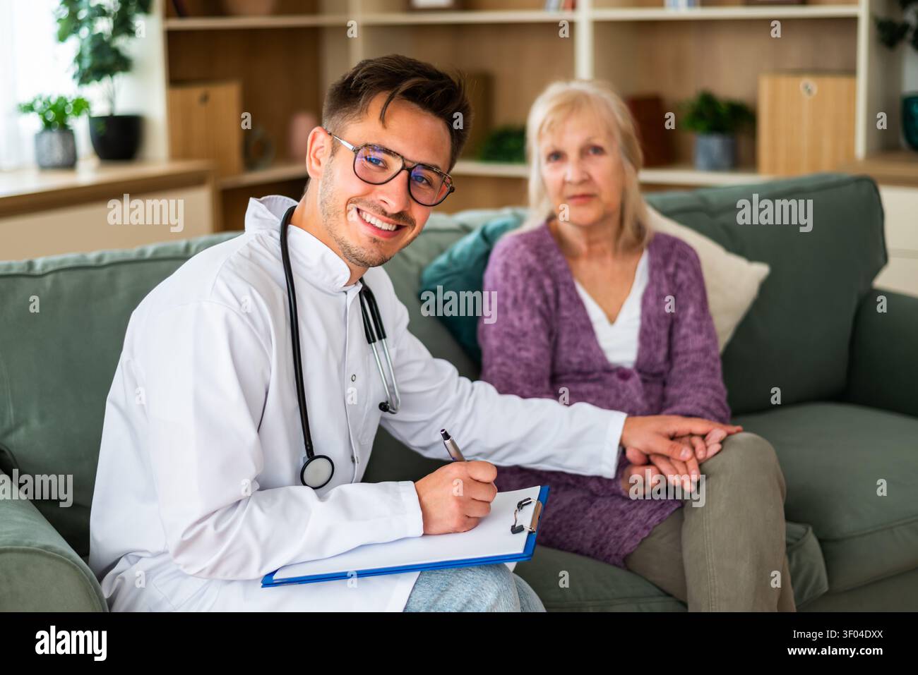 Un operatore sanitario cordiale sorride mentre prende appunti durante una visita a casa, tenendo delicatamente la mano di un paziente anziano per rassicurare e. Foto Stock