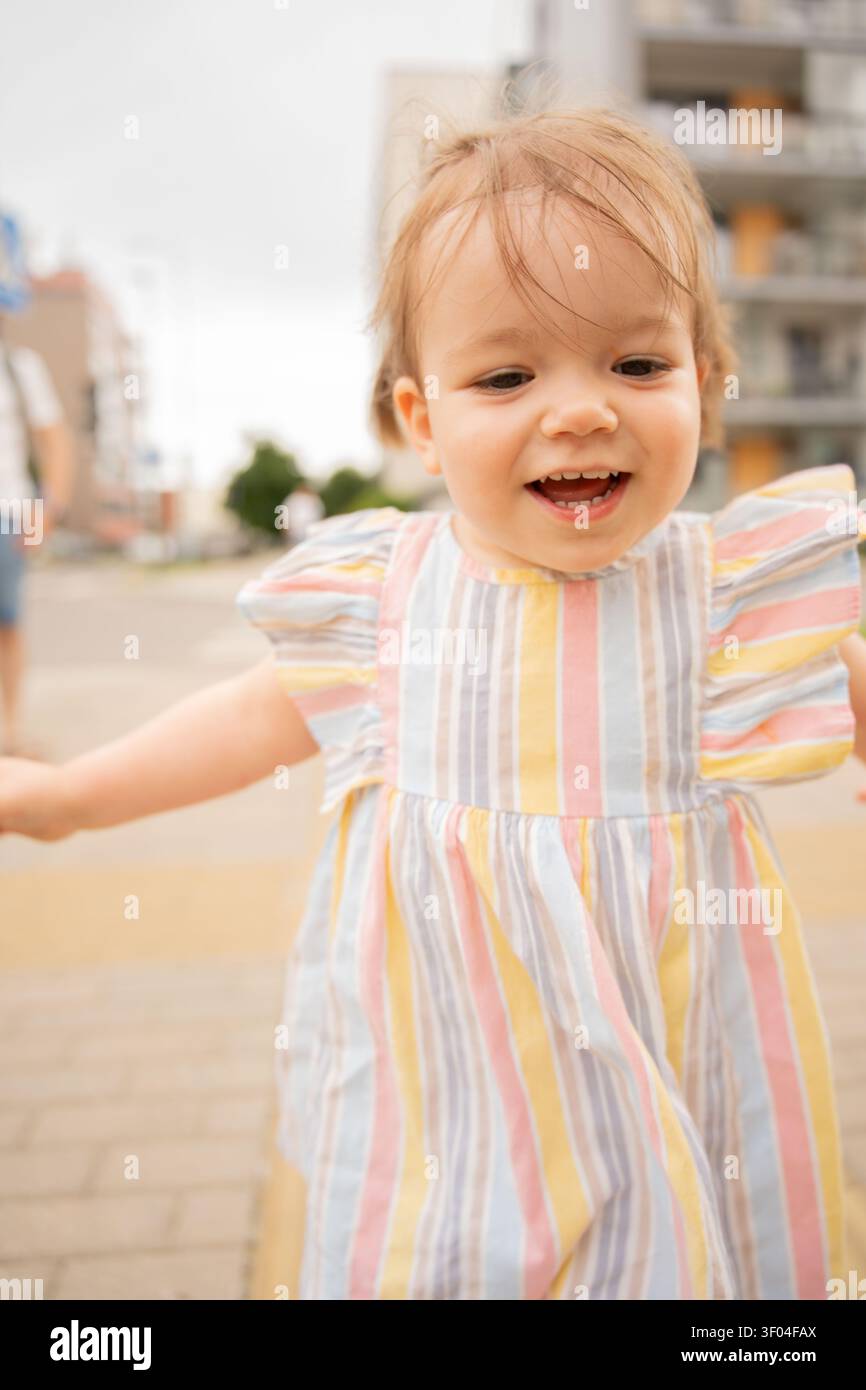 primo piano, ritratto schietto di un bambino sorridente che indossa un abito a righe pastello, capelli traforati dal vento e risate a bocca aperta catturate su un marciapiede urbano Foto Stock