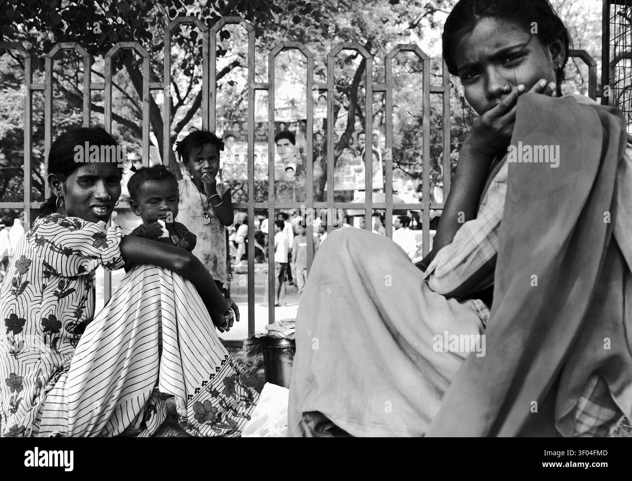 Ritratto documentaristico in bianco e nero di due donne indiane sedute con bambini vicino a una recinzione, con un cimitero sullo sfondo. Strada autentica Foto Stock
