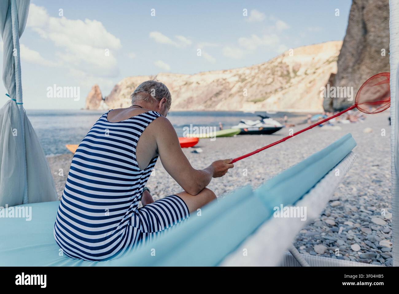 L'uomo in spiaggia indossa un costume da bagno a righe e siede su una poltrona reggendo una rete rossa a farfalla su una costa rocciosa Foto Stock