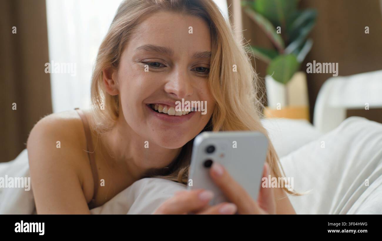 Una bella donna caucasica che si sveglia a letto riceve un messaggio sul telefono cellulare che chatta con un sms dell'app online sorridente che si trova in camera la mattina Foto Stock