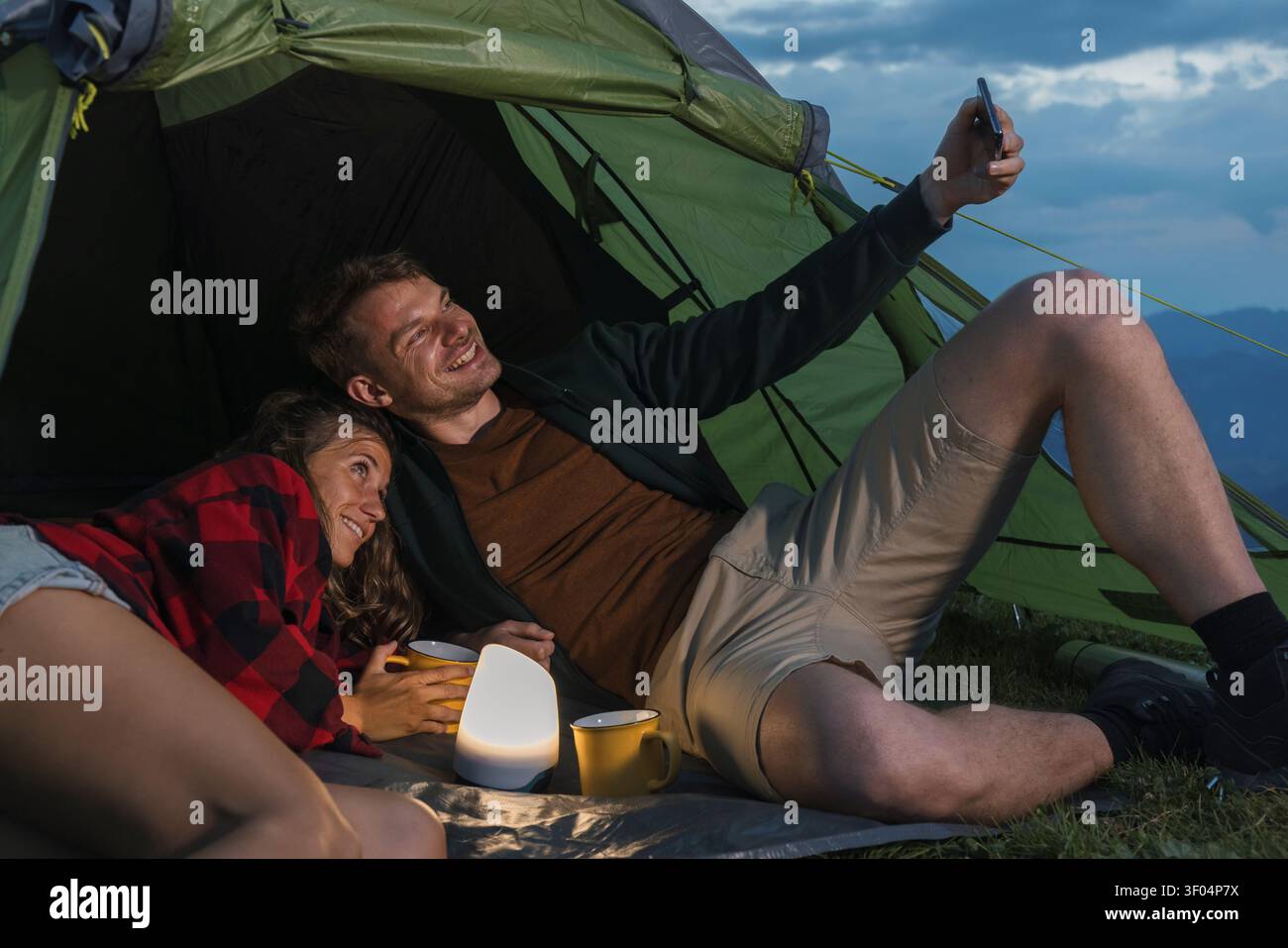 Due escursionisti si rilassano nella loro tenda al crepuscolo dopo una giornata di trekking in montagna. Sorridono mentre fanno un selfie insieme. Foto Stock