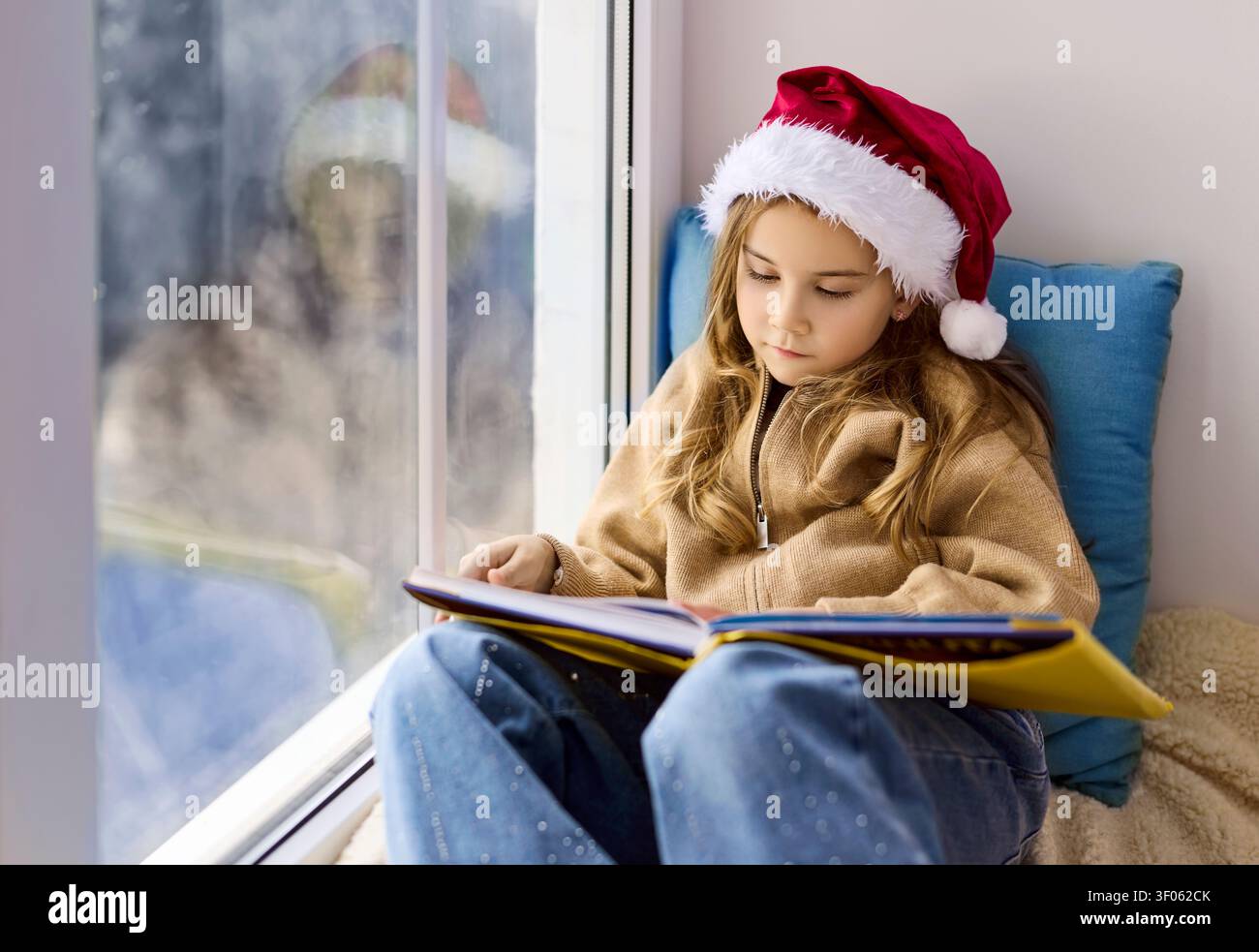 Natale bambino carino nel cappello di Babbo Natale che legge un libro alla finestra, seduto sul davanzale Foto Stock