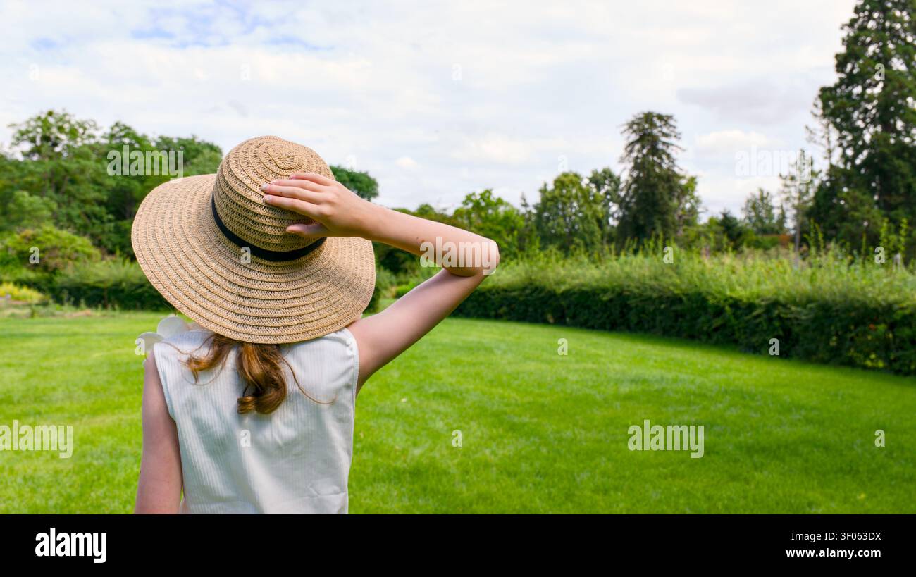 Una giovane donna, vista di spalle, si trova in un vivace parco verde, tiene il suo cappello di paglia mentre guarda verso alberi lontani e un cielo nuvoloso Foto Stock