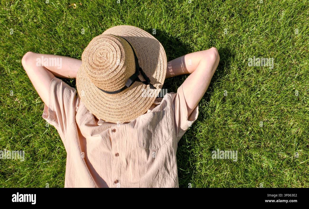 Da una vista aerea, una persona giace direttamente sull’erba verde brillante con un cappello di paglia che copre il viso e le mani strette dietro la testa. Foto Stock