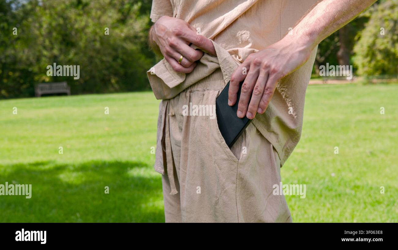 Una persona, vestita con abiti casual di colore chiaro, mette uno smartphone nero nella tasca dei pantaloni mentre è in piedi in un parco soleggiato. Foto Stock