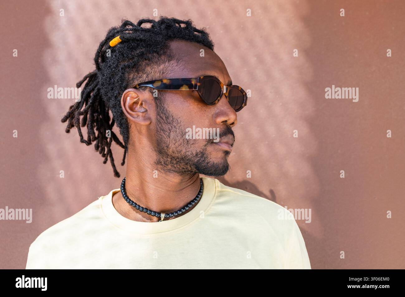 Ritratto laterale di un uomo cubano che indossa occhiali da sole, dreadlock e una collana con perline, appoggiato su una calda parete testurizzata con luce naturale. Foto Stock