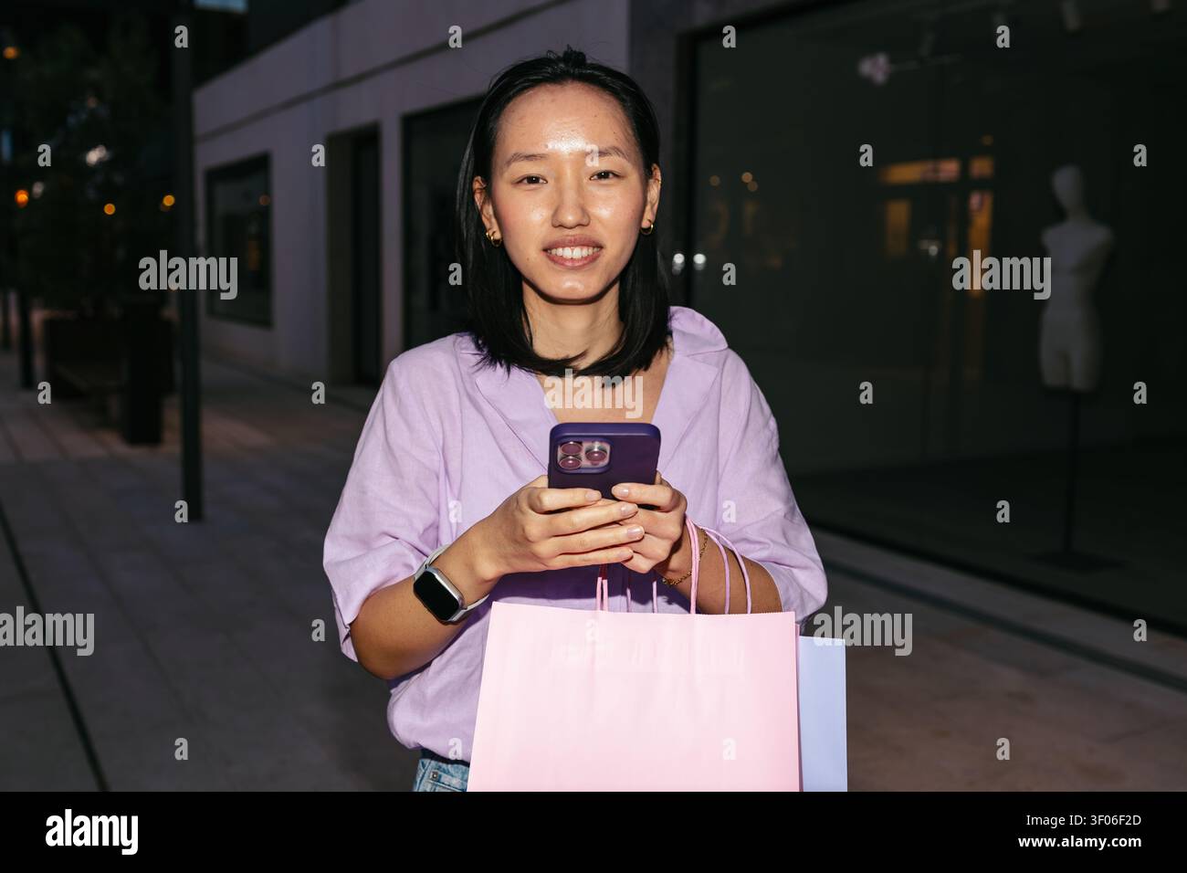 Una donna asiatica che tiene in mano le borse della spesa utilizza il suo smartphone in un ambiente urbano di notte, indossando un top casual in lavanda e uno smartwatch, incarnando un moder Foto Stock