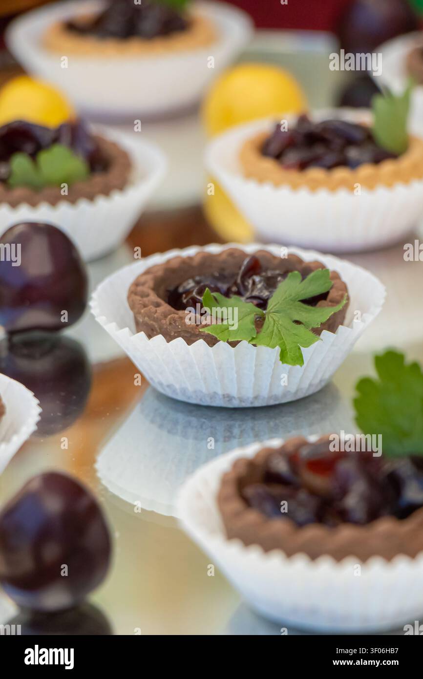 Close-up di tartlette al cioccolato artigianali ripiene di marmellata di ciliegie dolce, disposte su un elegante esposizione di dessert con messa a fuoco selettiva, ideale per b Foto Stock