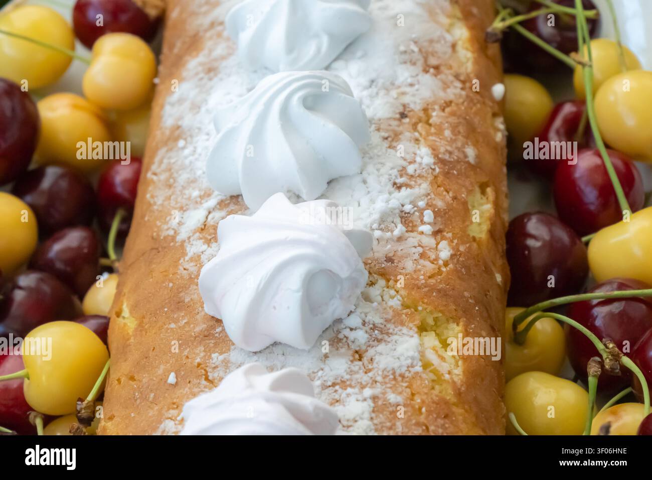 Torta casalinga a forma di pane ricoperta di meringhe bianche e zucchero a velo, circondata da ciliegie rosse e gialle per un nature morto rustico di dessert estivo. Foto Stock