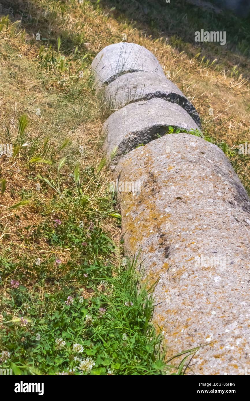 Cilindri di pietra erosi distesi nell'erba con muschio e piante selvatiche, che mostrano un dettaglio archeologico rustico del vecchio patrimonio rurale e del decadimento naturale. Foto Stock