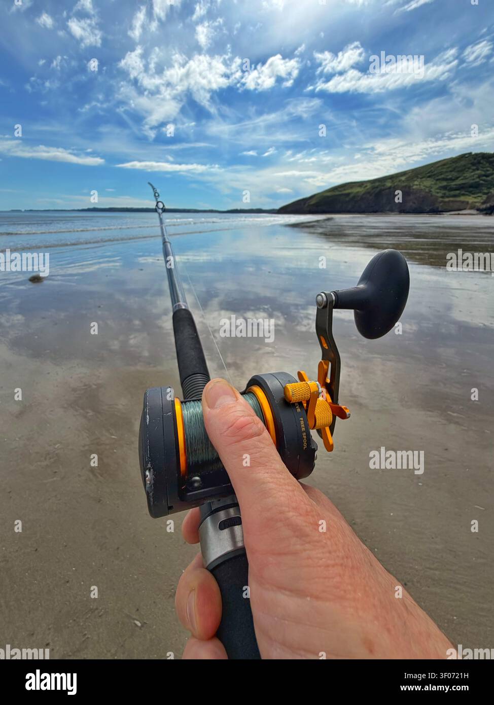 Mano di un uomo che controlla un mulinello a moltiplicatore durante la pesca in mare su una spiaggia gallese, Galles, Regno Unito Foto Stock