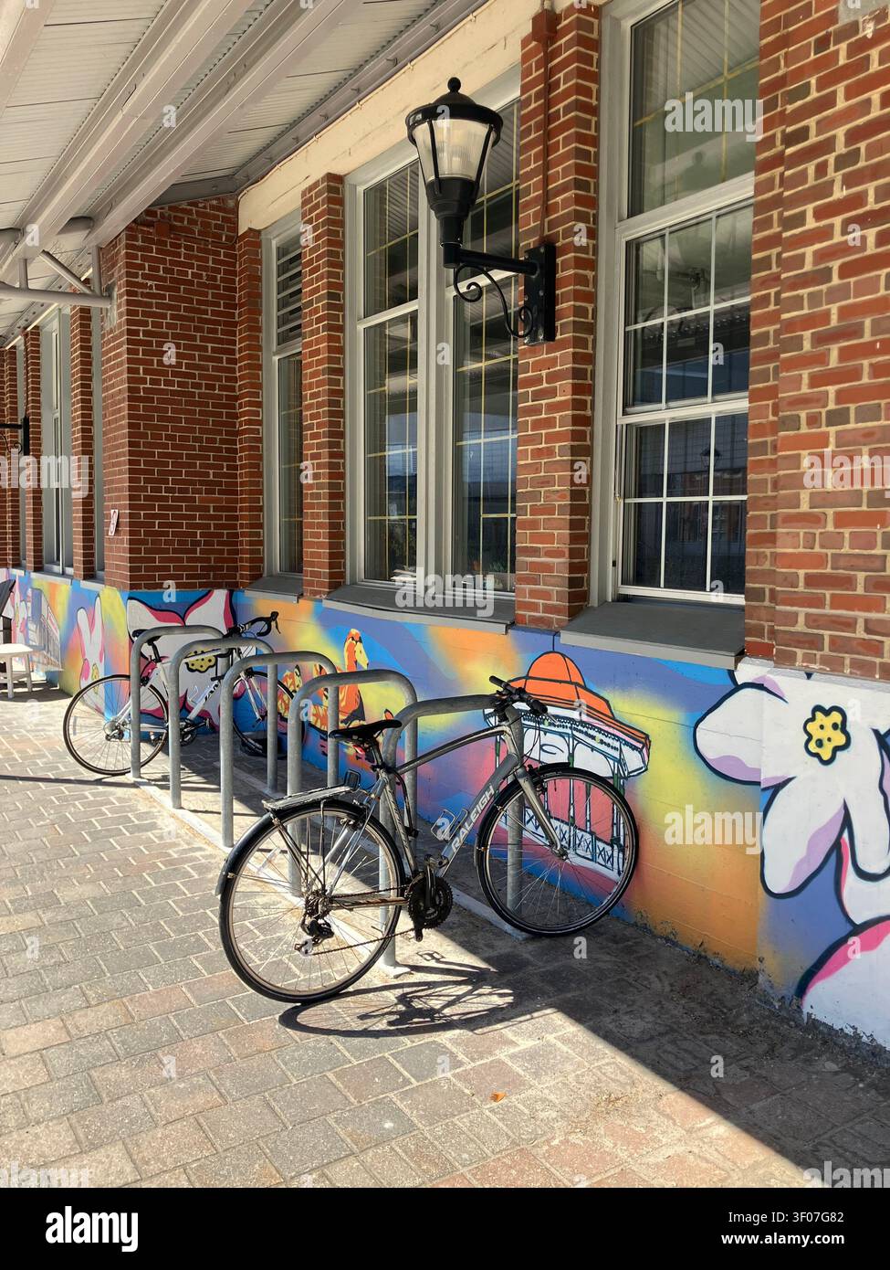 Biciclette parcheggiate in rastrelliere metalliche davanti a un edificio in mattoni con murale colorato e lampione classico. Distretto del porto di Halifax. - Immagine stock catturata con smartphone