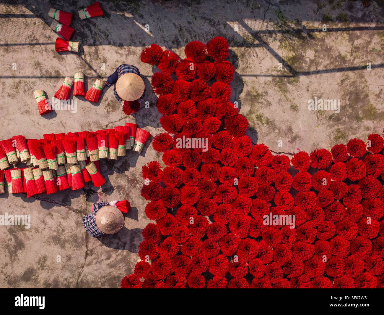 Vista aerea dei lavoratori che indossano cappelli conici che sistemano fasci di bastoncini di incenso rosso brillante per asciugare su una superficie di cemento in Vietnam. Foto Stock