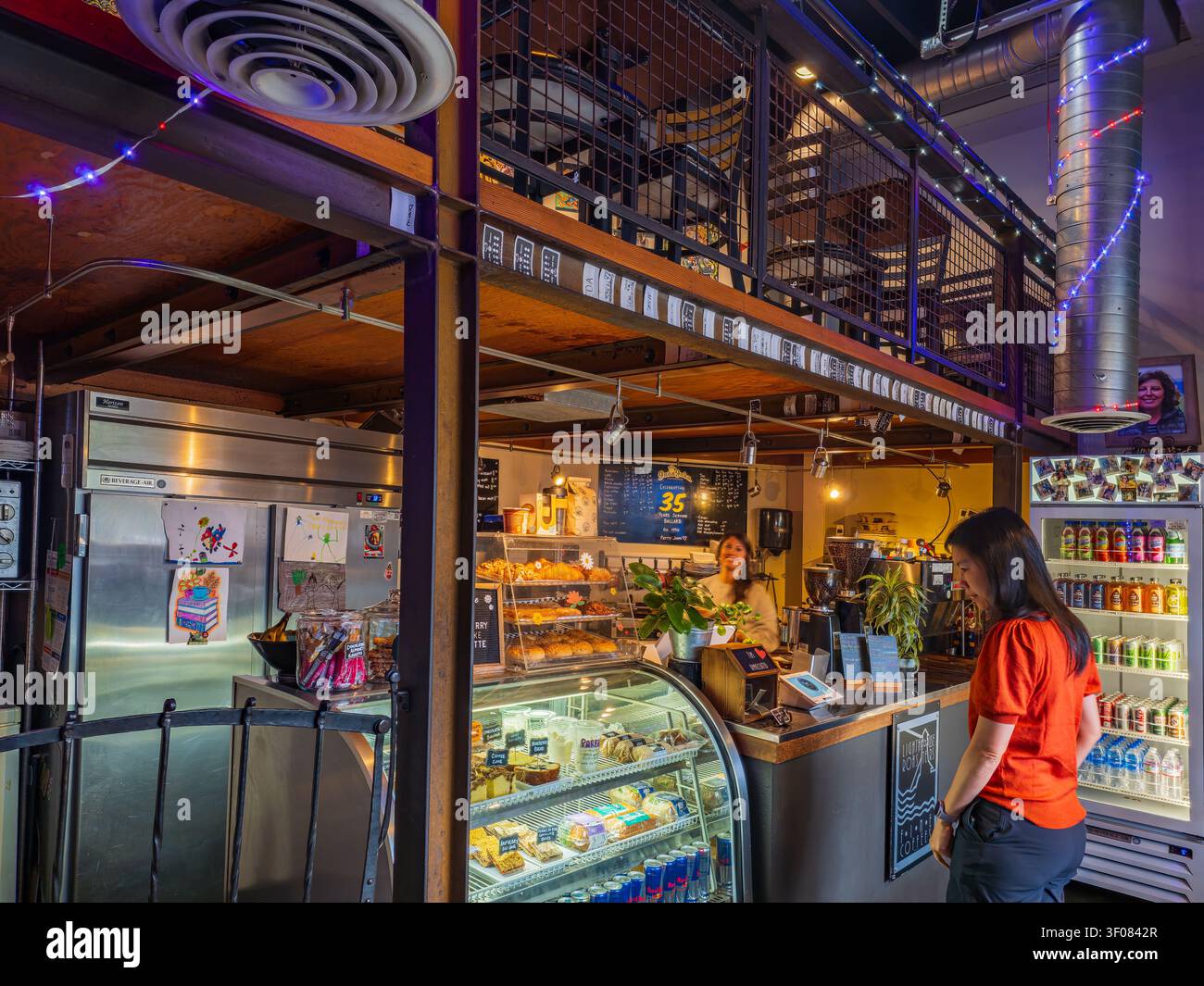 Stato di Washington, Stati Uniti, 26 GIU 2026 - Una donna con una camicia arancione è al bancone di un caffè moderno con un piano intermedio, sta ordinando Foto Stock