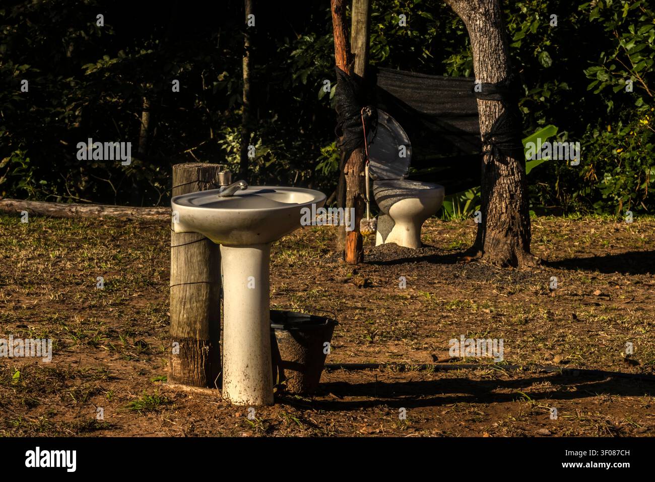 Un'installazione rustica da bagno all'aperto con un lavandino su piedistallo bianco e una vasca wc situata in un campo rurale circondato da alberi a Bra Foto Stock