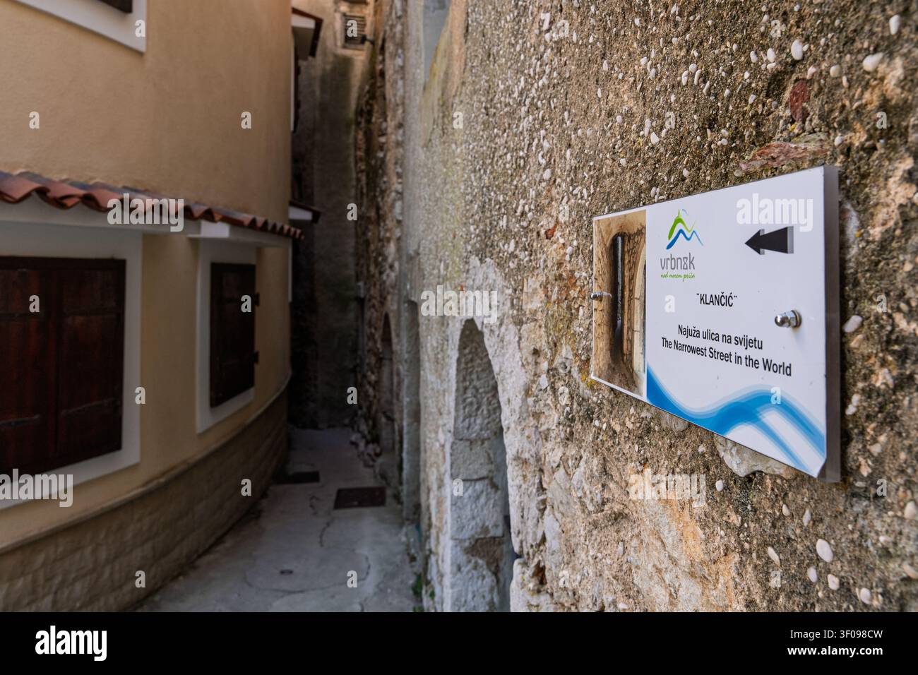 insegna stradale più stretta che indica la direzione in un vicolo urbano con pareti di cemento e mattoni, primo piano informativo, colori bianco e blu, design moderno Foto Stock