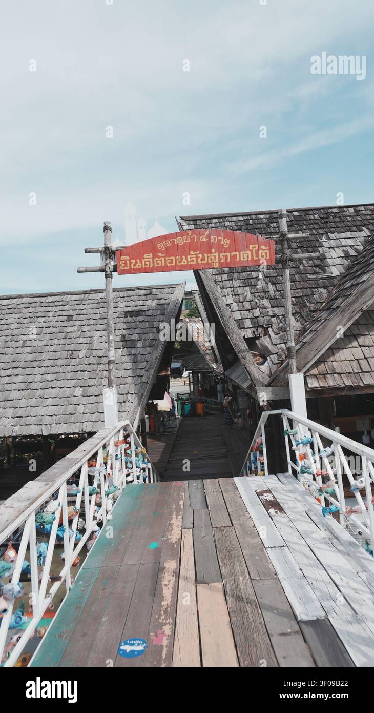 Ponte in assi di legno con ringhiera bianca, ingresso al mercato, cartello con scritta in thailandese, edifici in legno di paglia Foto Stock