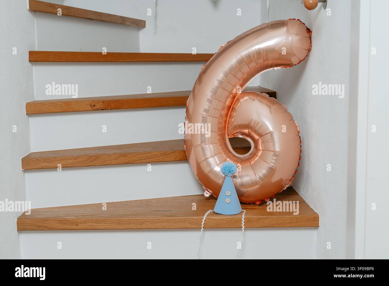 Un palloncino con il numero sei poggia su una scala di legno, adornato con un cappello da festa blu, celebrando una festa di compleanno in un ambiente interno con pareti bianche Foto Stock