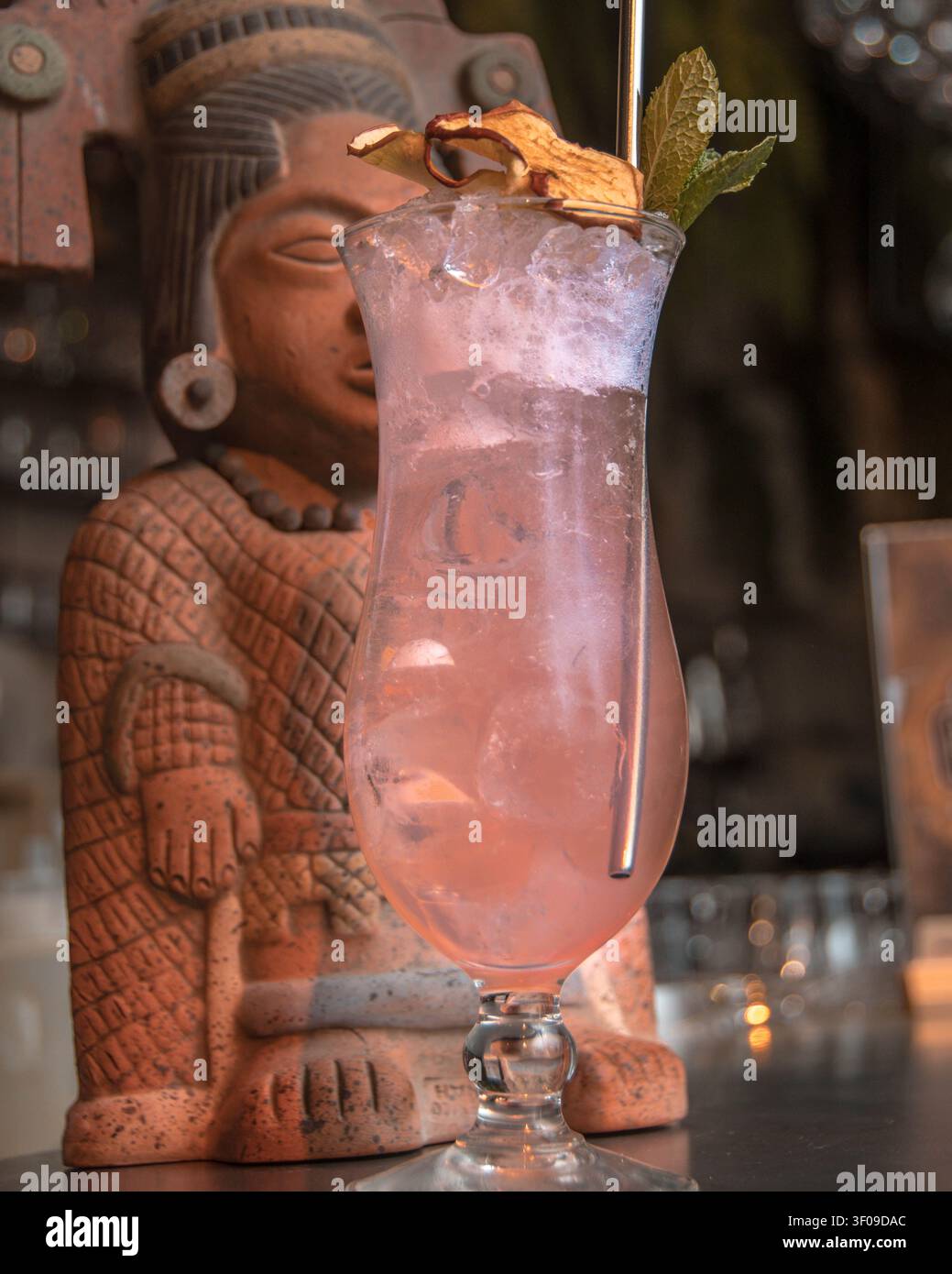 Cocktail tropicale rosa con menta e agrumi essiccati servito accanto a una statua tiki Foto Stock