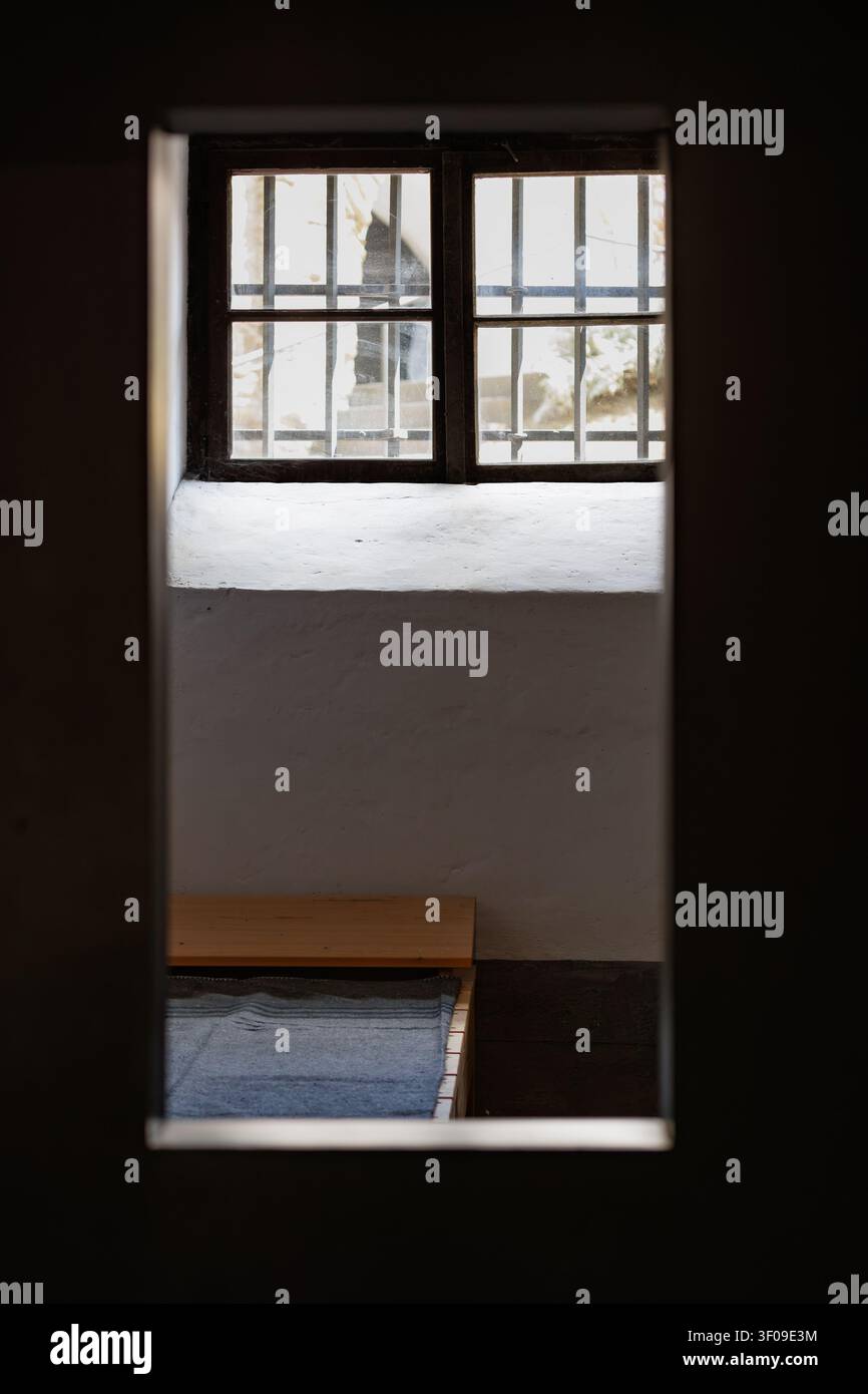 Interno storico di una cella di prigione visto attraverso la porta con finestra sbarrata e letto Foto Stock