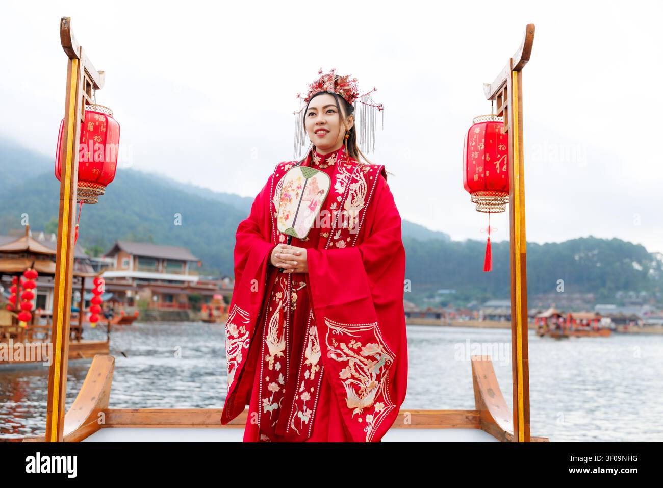 Una donna in un ornato hanfu cinese tradizionale rosso tiene un ventaglio manuale mentre è in piedi su una barca di legno nel villaggio di Ban Rak Thai. Foto Stock