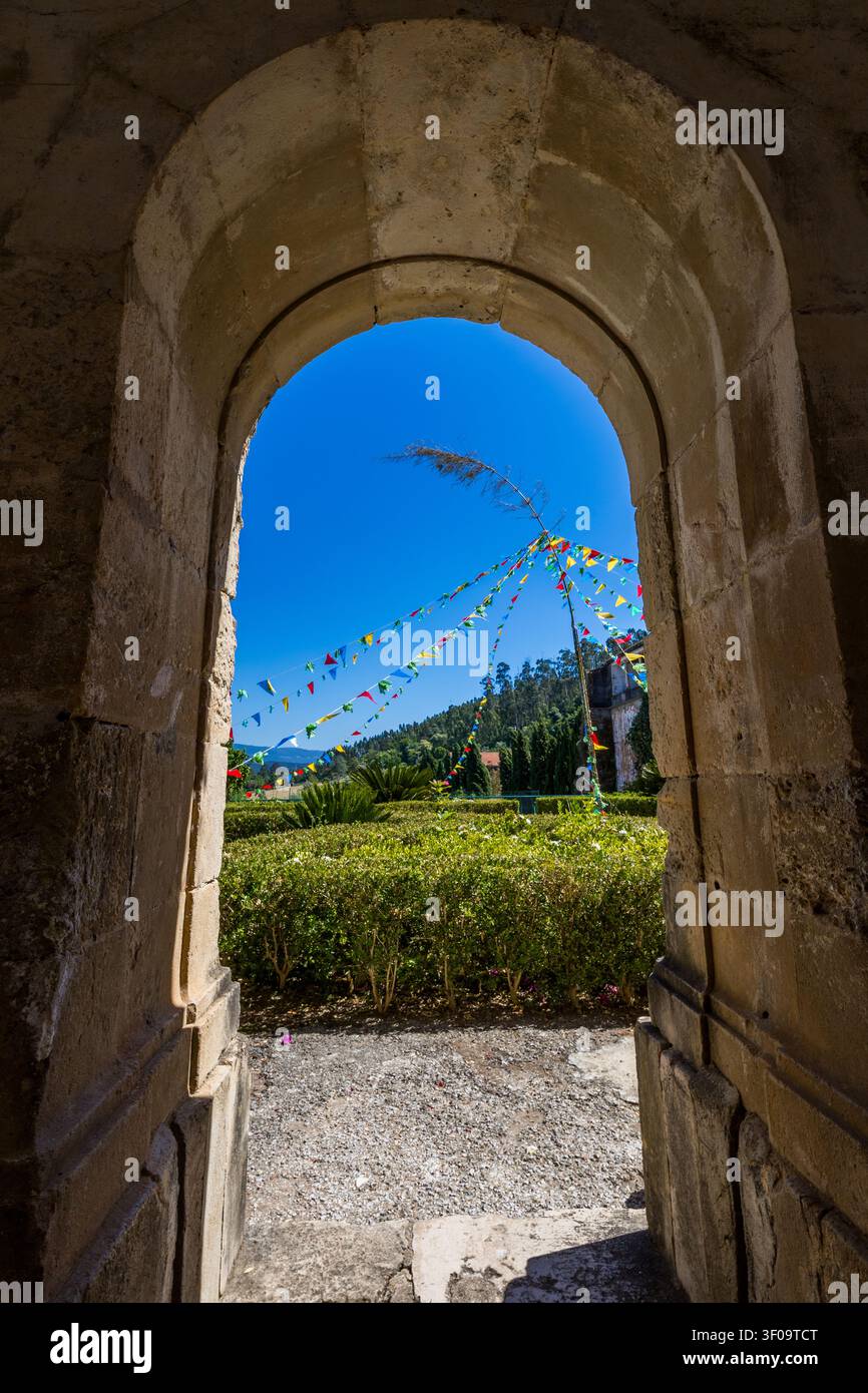 Incorniciato da un arco di pietra storico, un giardino cortile illuminato dal sole in Portogallo mostra bandierine colorate per le feste contro un cielo blu terso. Foto Stock