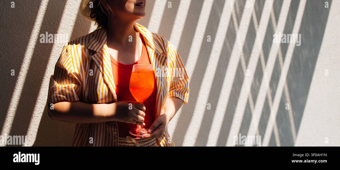 Donna in piedi contro un muro con ombre diagonali, tiene un bicchiere di bevanda arancione, vestita con una camicia a righe e un top rosso in uno spazio interno ben illuminato Foto Stock