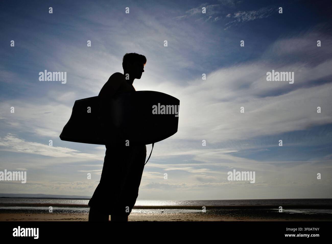 Silhouette di un giovane uomo con bodyboard in piedi su una spiaggia che guarda verso il mare Foto Stock