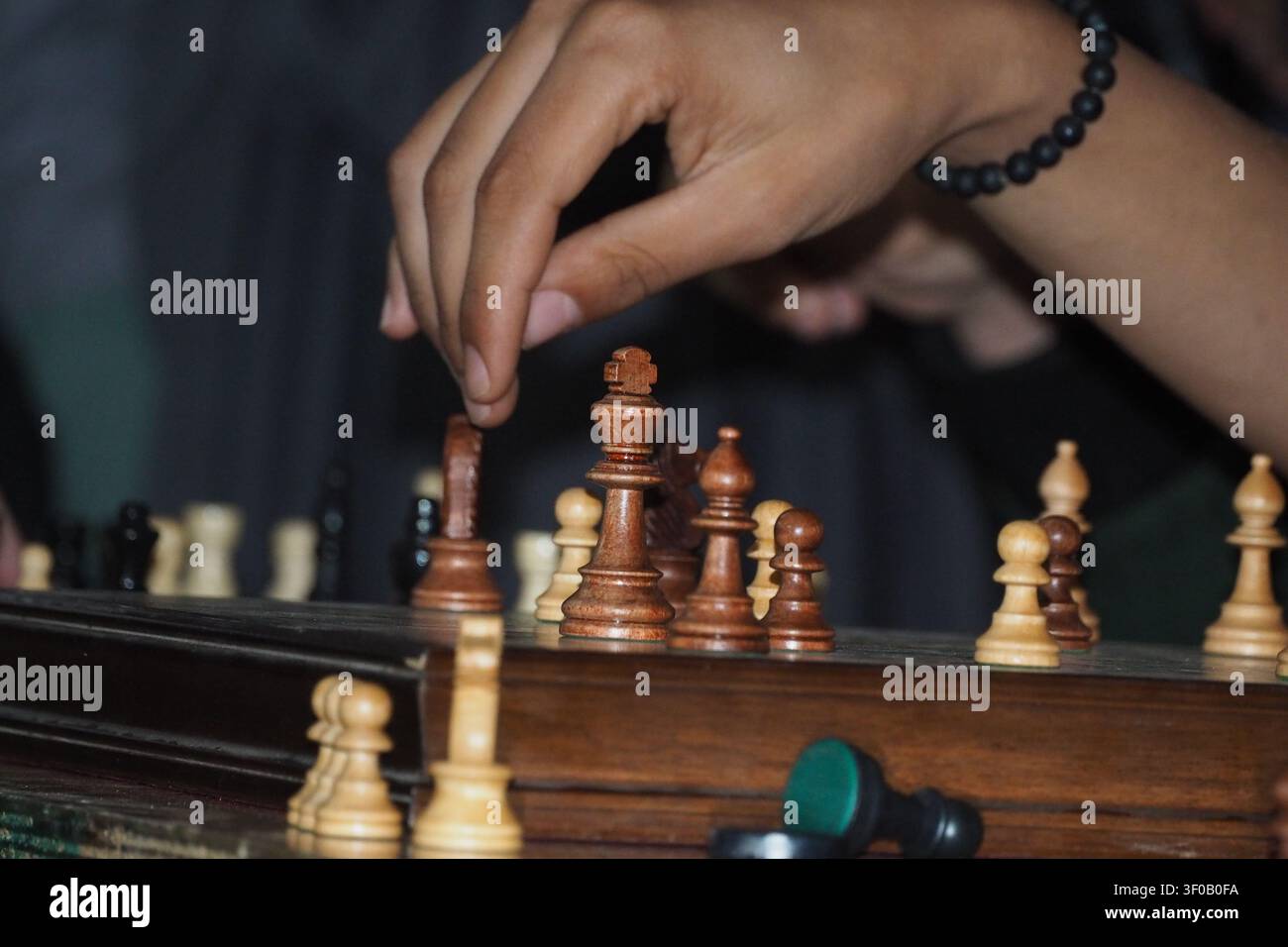 Uno scatto mirato che mostra la mano di una persona con un braccialetto che raccoglie o sposta con attenzione una pedina re/regina su una classica scacchiera di legno. Foto Stock