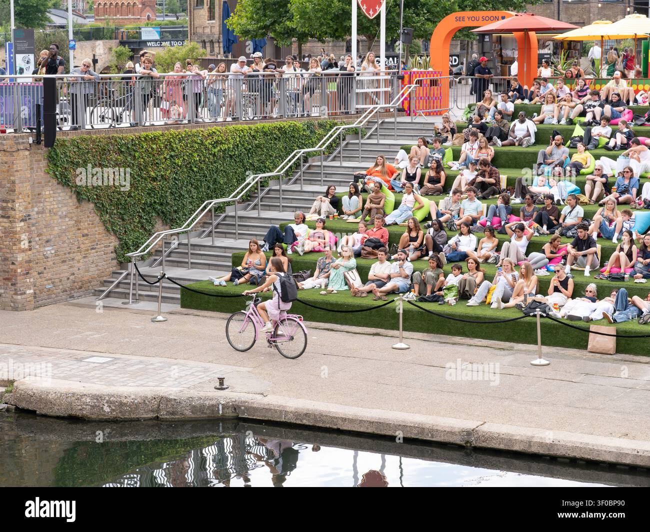 Londra, Regno Unito – 9 luglio 2025: Persone che si rilassano sui gradini verdi del lato del canale di Londra, seduta sull’erba vicino a un canale, una persona che passa in bicicletta, godendosi un cas Foto Stock