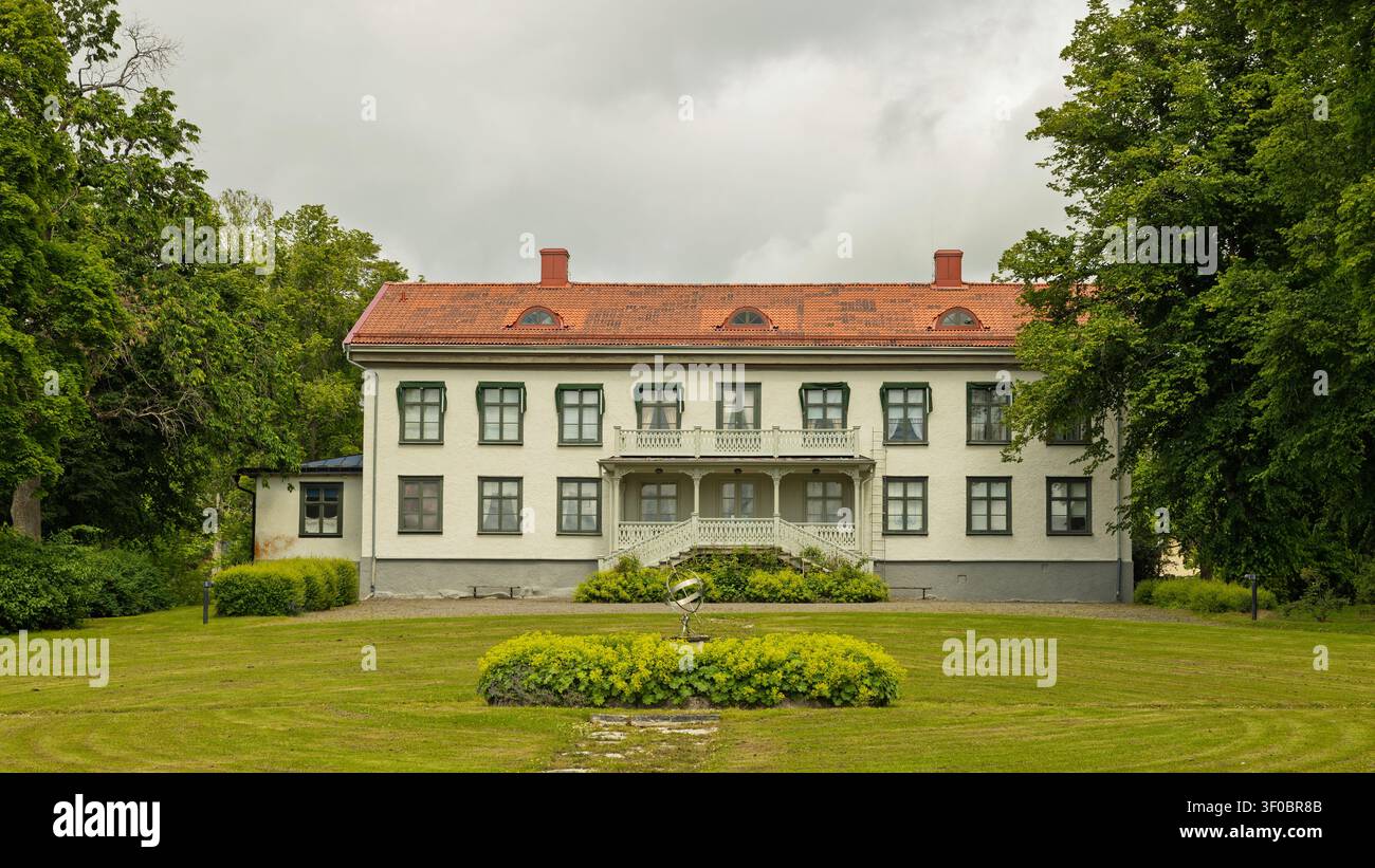 Karlskoga, Svezia - 18.06.2026: Una grande tenuta a due piani caratterizzata da una facciata bianca, tetto in terracotta e balcone ornato in un giardino ben curato Foto Stock