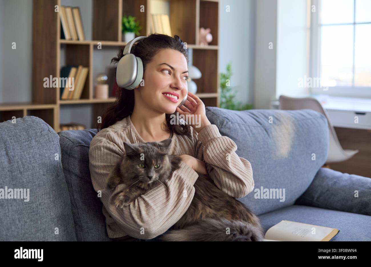 Giovane donna felice con le cuffie che tiene un gatto sul divano, rilassandosi a casa Foto Stock